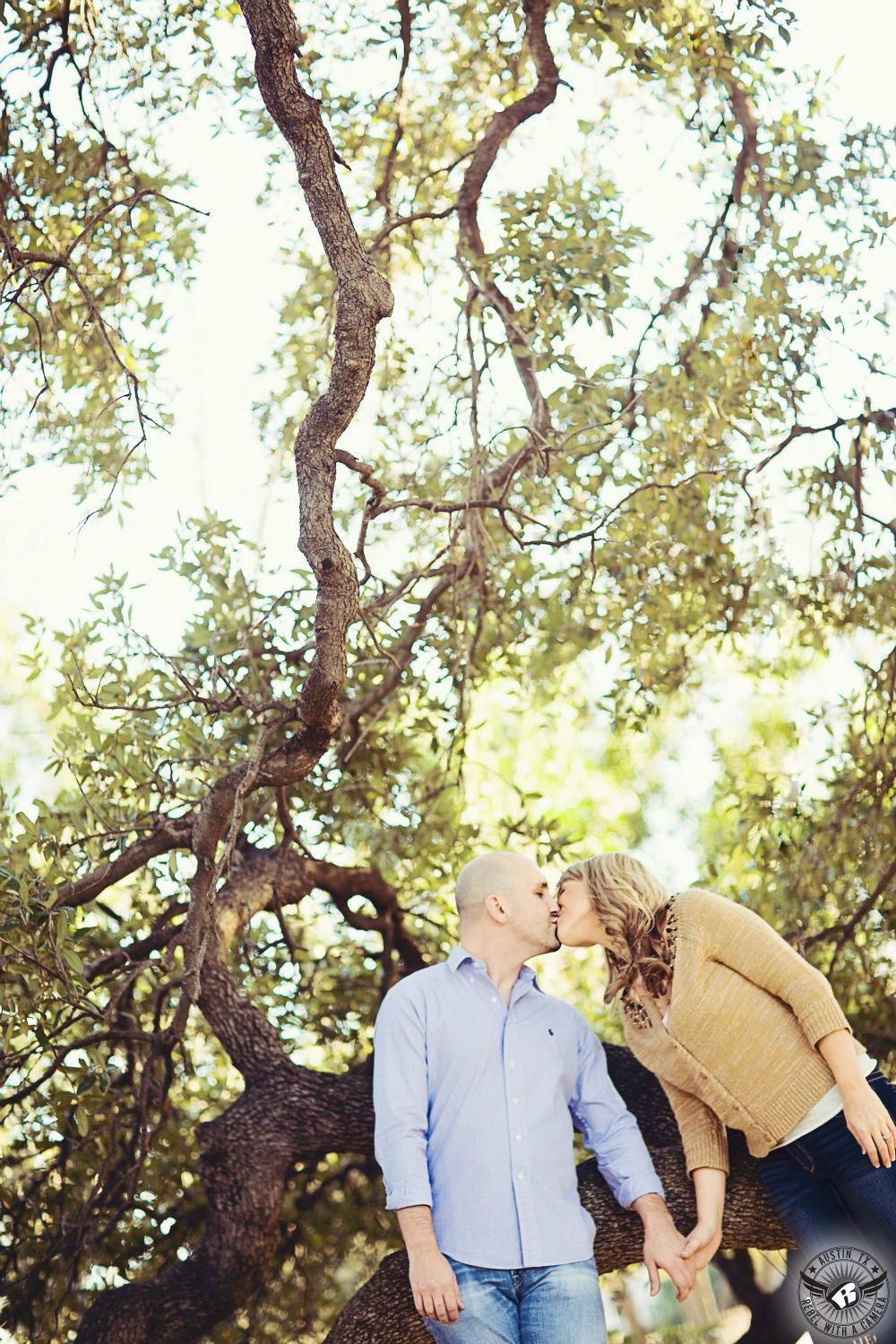 Curly blond woman in a tan button up sweater and blue jeans leans in and kisses guy wearing a light blue button up dress shirt and washed out blue jeans while holding hands in front of scattered tree branches, leaves and the bright sky in this fanciful engagement image at the Capitol of Texas in Austin.